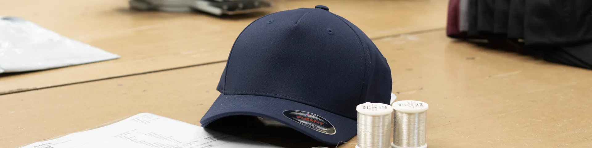 A navy cap on the bench at BR Apparel in Bradford, waiting to be embroidered, with two cones of silver thread and the job sheet beside it